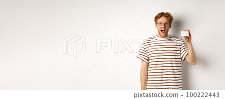 Shopping and finance concept. Amazed young man with beard and red hair showing plastic credit card and looking impressed, white background Shopping and finance concept. Amazed young man with beard and red hair showing plastic credit card and looking impressed, white background 100222443