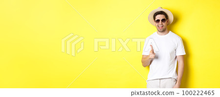 Confident and cheeky guy on vacation flirting with you, pointing finger at camera and winking, wearing summer hat with sunglasses, yellow background 100222465