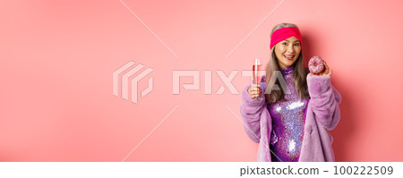 Beautiful and stylish asian senior woman having fun, drinking champagne and eating sweet donut, smiling at camera, standing over pink background 100222509