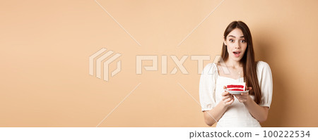Excited birthday girl looking amazed, holding cake with candle, making b-day wish, standing on beige background Excited birthday girl looking amazed, holding cake with candle, making b-day wish, standing on beige background 100222534