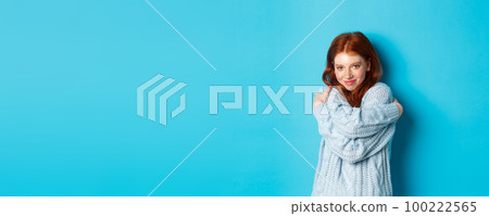 Happy cute redhead girl hugging herself, wearing comfortable and warm sweater, smiling at camera, standing over blue background 100222565