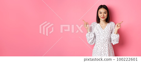 Beautiful asian woman making choice on shopping, pointing fingers sideways and showing variants, smiling at camera, standing over pink background 100222601