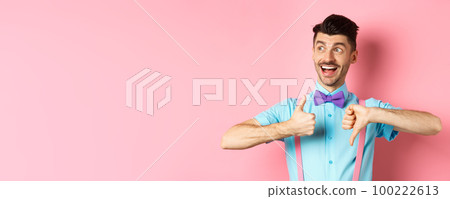 Cheerful man looking happy left, showing thumbs up down, judging product, give positive and negative feedback, standing over pink background Cheerful man looking happy left, showing thumbs up down, judging product, give positive and negative feedback, standing over pink background 100222613
