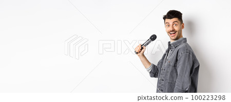 Profile of handsome smiling man holding mic, turn head at camera with excited face, singing karaoke and perform standup, standing on white background Profile of handsome smiling man holding mic, turn head at camera with excited face, singing karaoke and perform standup, standing on white background 100223298