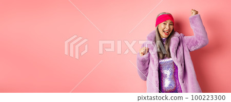 Shopping concept. Cheerful senior asian woman dancing and having fun, wearing stylish purple faux fur coat and dress, winning prize and smiling, standing over pink background Shopping concept. Cheerful senior asian woman dancing and having fun, wearing stylish purple faux fur coat and dress, winning prize and smiling, standing over pink background 100223300