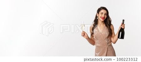 Winter holidays celebration concept. Beautiful woman enjoying Christmas and New Year party, drinking champagne and winking at camera, standing in elegant dress 100223312