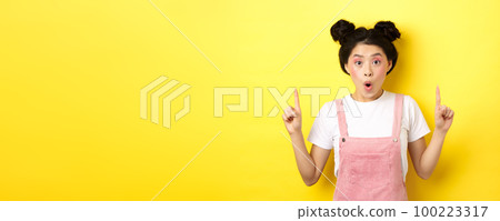 Excited asian female model with glamour makeup, pointing fingers up and say wow amazed, checking out promo deal, standing on yellow background 100223317
