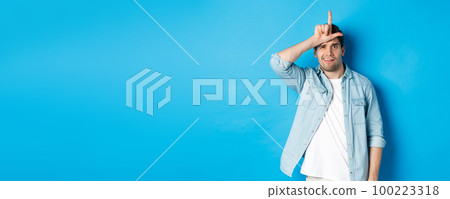 Rude guy mocking person for losing, showing loser sign on forehead and looking with dismay, standing against blue background 100223318