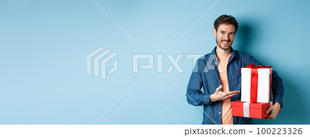 Young man in casual clothes buying romantic gifts on Valentines day, pointing at presents boxes and smiling, standing over blue background Young man in casual clothes buying romantic gifts on Valentines day, pointing at presents boxes and smiling, standing over blue background 100223326