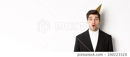 Close-up of attractive man in party hat and trendy suit, looking surprised, celebrating new year, standing against white background Close-up of attractive man in party hat and trendy suit, looking surprised, celebrating new year, standing against white background 100223328