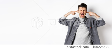 Annoyed guy shut ears and screaming, displeased by loud noise, yelling to stop noisy music, standing on white background 100223329