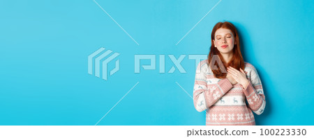 Image of dreamy redhead girl close eyes and holding hands on heart, feeling nostalgic, remember or imagine something, standing over blue background Image of dreamy redhead girl close eyes and holding hands on heart, feeling nostalgic, remember or imagine something, standing over blue background 100223330