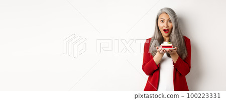 Surprised asian middle-aged woman holding piece of tasty cake, looking amazed at camera, standing over white background Surprised asian middle-aged woman holding piece of tasty cake, looking amazed at camera, standing over white background 100223331