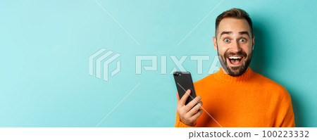 Close-up of happy handsome man writing message on mobile phone, holding smartphone and smiling, standing against turquoise background 100223332
