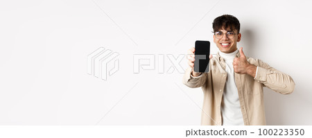 Portrait of handsome young man in glasses, showing empty smartphone screen and thumb up, recommending online app or store, standing pleased on white background Portrait of handsome young man in glasses, showing empty smartphone screen and thumb up, recommending online app or store, standing pleased on white background 100223350