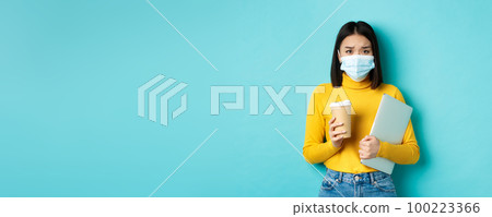 Covid-19, social distancing and pandemic concept. Worried asian woman in medical mask, frowning sad, holding laptop for work and cup of coffee Covid-19, social distancing and pandemic concept. Worried asian woman in medical mask, frowning sad, holding laptop for work and cup of coffee 100223366
