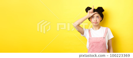 Sad girl showing loser sign on forehead and sulking upset, feeling disappointed in herself, standing on yellow background 100223369
