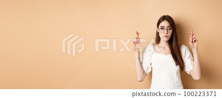 Hopeful girl in glasses cross fingers for good luck, biting lip nervous and looking at camera, waiting for results, praying or making wish, beige background 100223371