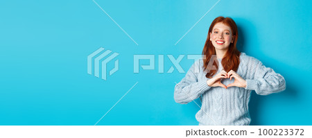 Cute redhead girl in sweater showing heart sign, I love you gesture, smiling at camera, standing against blue background 100223372