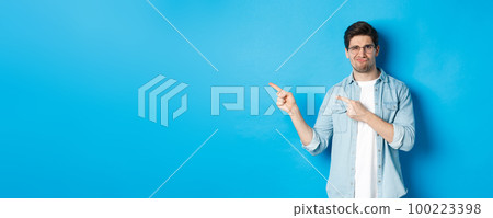 Displeased and skeptical male model in glasses, pointing fingers left at something bad, showing awful advertisement, standing over blue background 100223398
