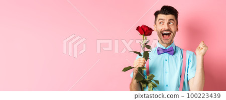 Romance and Valentines day concept. Cheerful man in bow-tie screaming from happiness, holding red rose and jumping on date, standing over pink background 100223439