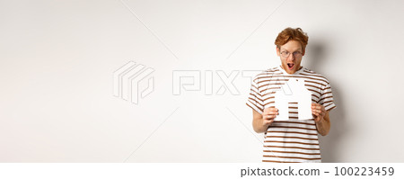 Amazed young man with red hair, staring at paper house cutout with excitement, standing over white background Amazed young man with red hair, staring at paper house cutout with excitement, standing over white background 100223459
