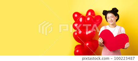 Happy Valentines day. Silly and beautiful asian woman smiling dreamy, showing red heart, imaging romantic date with lover, standing on yellow background Happy Valentines day. Silly and beautiful asian woman smiling dreamy, showing red heart, imaging romantic date with lover, standing on yellow background 100223487
