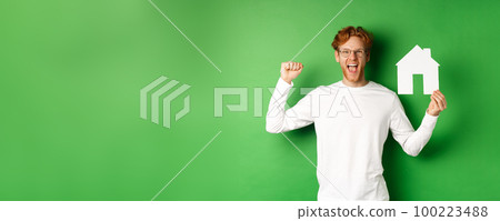 Real estate. Happy caucasian man in glasses with red hair celebrating, showing paper house cutout and making fist pump sign, shouting for joy, buying property, green background 100223488
