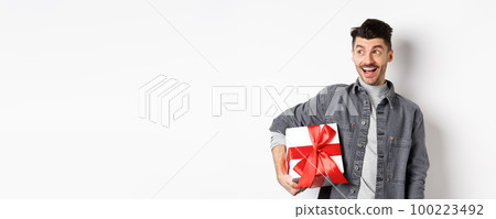 Cheerful handsome guy looking at empty space with amazed smile, checking out special valentines day offer, holding big romantic gift box, standing on white background 100223492