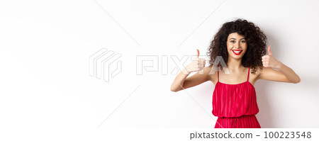 Attractive young woman recommending promo offer, showing thumb up and smiling, like product, standing in festive red dress on white background Attractive young woman recommending promo offer, showing thumb up and smiling, like product, standing in festive red dress on white background 100223548