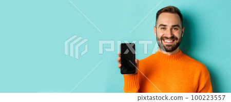 Close-up of handsome bearded guy in orange sweater, showing smartphone screen and smiling, showing promo online, turquoise background Close-up of handsome bearded guy in orange sweater, showing smartphone screen and smiling, showing promo online, turquoise background 100223557