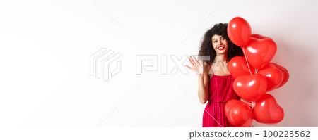 Beautiful lady with curly hair, standing near valentines holiday balloons and waving hand, saying hello, standing against white background 100223562