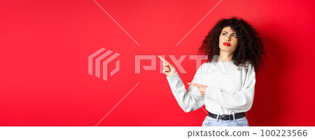 Skeptical frowning girl with curly hair, pointing and looking left hesitant, standing upset on red background Skeptical frowning girl with curly hair, pointing and looking left hesitant, standing upset on red background 100223566