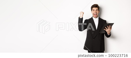 Portrait of excited handsome businessman in black suit, holding clipboard and making fist pump, achieve goal and rejoicing, standing over white background 100223716