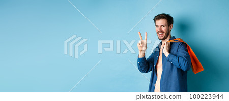Cheerful guy holding orange shopping bag on shoulder, smiling and showing peace sign, standing on blue background 100223944