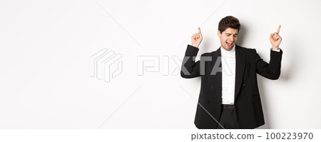 Image of successful handsome businessman dancing, enjoying party, pointing fingers up and having fun, standing over white background, wearing black suit 100223970