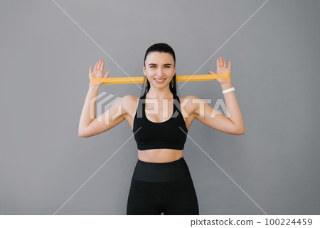 A young smiling strong athletic fitness trainer, a female instructor in a black tracksuit is spending time in the home gym with a fitness elastic band on the back muscles. The concept of sports A young smiling strong athletic fitness trainer, a female instructor in a black tracksuit is spending time in the home gym with a fitness elastic band on the back muscles. The concept of sports 100224459