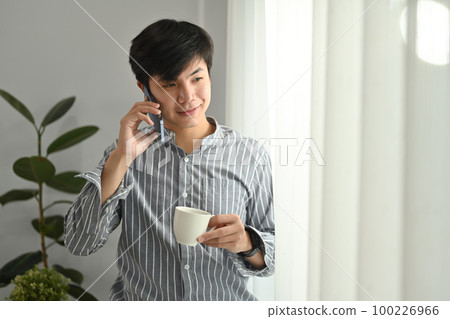 Portrait of smiling asian businessman holding coffee cup and talking on mobile phone at bright home office Portrait of smiling asian businessman holding coffee cup and talking on mobile phone at bright home office 100226966