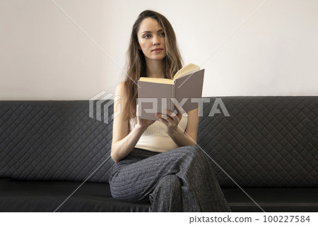 Beautiful, brooding young woman sits on the couch, holds an open book, and looks away Beautiful, brooding young woman sits on the couch, holds an open book, and looks away 100227584