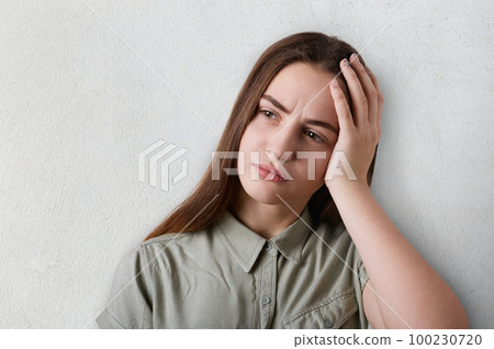 A portrait of thoughtful girl with long dark hair and big brown eyes wearing grey T-shirt having a sad expression holding her hand on her head. A tired girl having deep thoughts about something 100230720