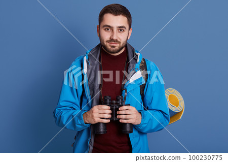 Picture of delighted glad man having pleasant facial expression, holding binoculars in both hands, looking directly at camera, having sleeping pad aback, wearing clothes for camping. Trip concept. 100230775