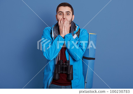 Image of impressed confused handsome explorer covering his mouth with both hands, looking directly at camera, wearing blue jacket, dark blue sweatshirt, having all equipment for hiking alone. 100230783