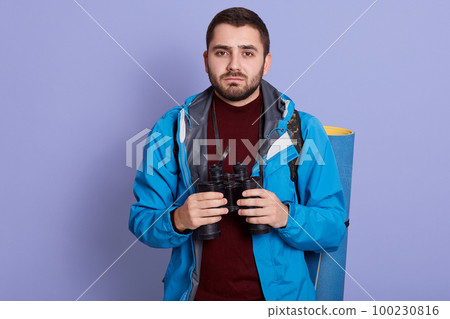 Horizontal shot of young handsome tourist man with beard, wearing blue jacket and sweater, using binoculars with confident and serious expression. Backpacking, traveling, vacation, resting cpncept. 100230816