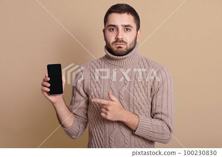 Close up portrait of nice calm handsome young man holding smartphone in one hand, pointing at it with forefinger, being upset, standing isolated over beige background in studio. Choice concept. Close up portrait of nice calm handsome young man holding smartphone in one hand, pointing at it with forefinger, being upset, standing isolated over beige background in studio. Choice concept. 100230830