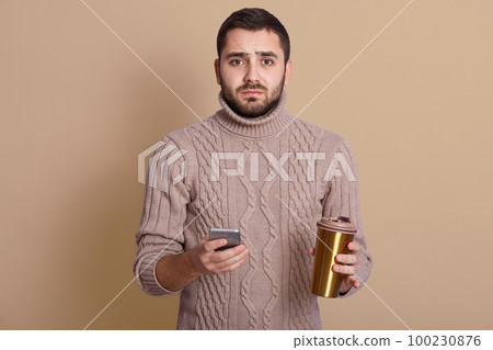 Studio shot of young Caucasian man using phone while holding thermo mug in hands, posing against beige background, wears sweater, drinking coffee or tea, looks at camera, having sad facial expression. 100230876