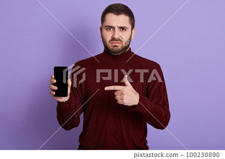 Horizontal shot of young sad man with disappointed facial expression, making gesture, holding smartphone, pointing at it with forefinger, being unsatisfied with choice and purchase. Tech concept. 100230890