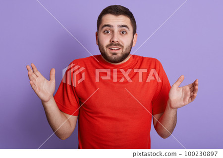 Close up portrait of young man posing isolated over purple background being at loss, showing helpless gesture with arm and hands, spreading his hands, wearing casual red t shirt. People concept. Close up portrait of young man posing isolated over purple background being at loss, showing helpless gesture with arm and hands, spreading his hands, wearing casual red t shirt. People concept. 100230897