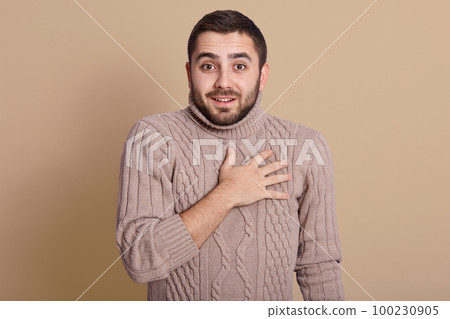 Indoor studio shot of positive energetic young man with black hair, wearing beige sweater, putting hand on chest, looking directly at camera, having peaceful facial expression. Emotions concept. 100230905