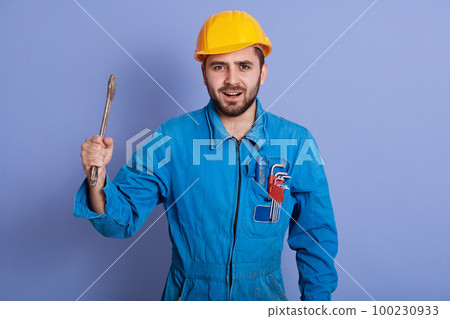 Half length portrait of emotional energetic young engineer opening mouth, looking directly at camera, working, holding metal wrench in one hand, standing isolated over blue background in studio. 100230933