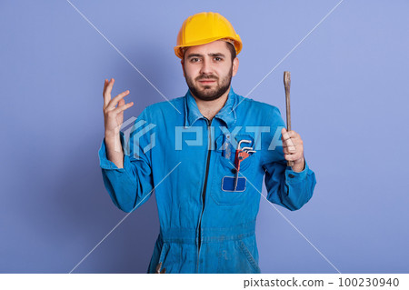 Puzzled male plumbers or car mechanic can`t understand where breakage is, shruging shoulders and raises hands in bewilderment, wearing yellow helmet and blue uniform, poses against sztudio wall. 100230940
